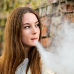 Vaping teenager. Young pretty white caucasian teenage girl with problem skin smoking an electronic cigarette the ruined brick wall on the street in the autumn. Bad habit.