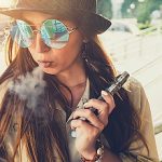 Pretty young hipster woman in black hat vape ecig, vaping device at the sunset. Toned image